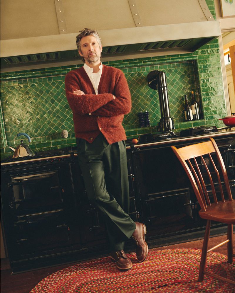 Man stands in kitchen, wearing cardigan and trousers, leaning against black range cooker with green tile backsplash and patterned rug.