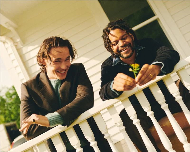 Two men, one holding a flower, smile on a porch with white railings. One man wears a brown blazer, while the other dons a black jacket.