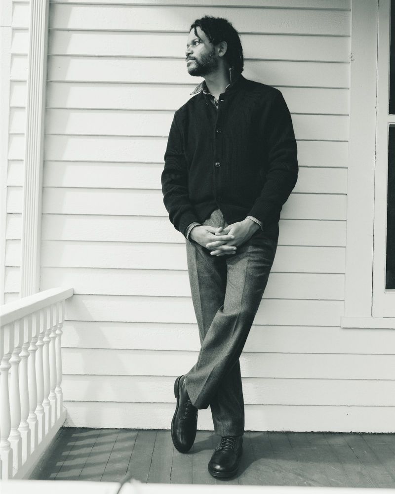 Portrait of a man with a beard, standing on a porch with a white wall and railing, wearing a cardigan and slacks, looking off to the side.