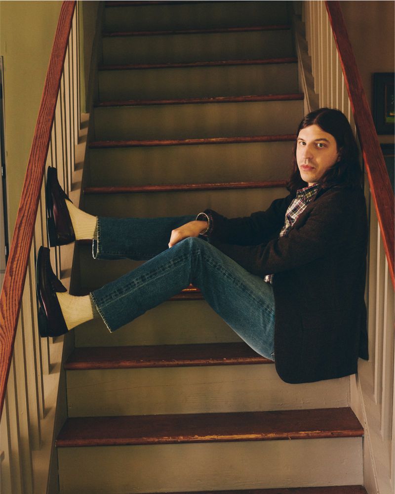Adult male with long hair and casual wear sitting on wooden stairs in a home with a plaid shirt and black jacket.