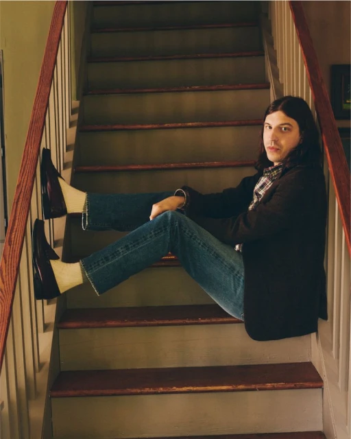 Adult male with long hair and casual wear sitting on wooden stairs in a home with a plaid shirt and black jacket.