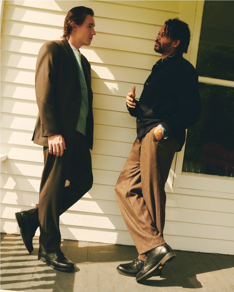 Two adult men in suits are standing outside a light beige house and talking during the day. One man is gesturing with his hand.