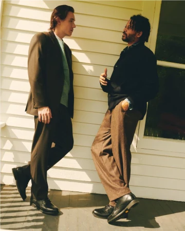 Two adult men in suits are standing outside a light beige house and talking during the day. One man is gesturing with his hand.