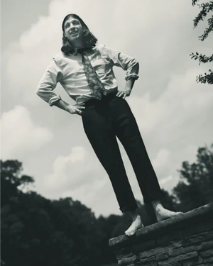 Monochrome low-angle portrait of a barefoot man in a button-down shirt and tie posing outdoors against an overcast sky.