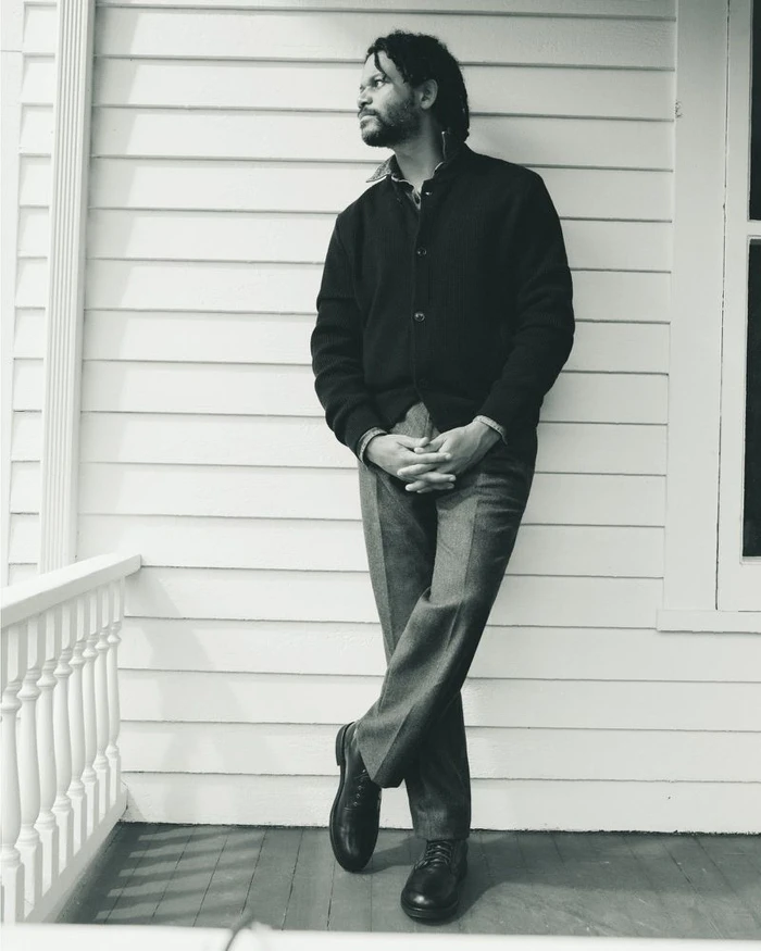 Man wearing a dark cardigan and trousers leaning against a white wooden paneled wall