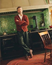 Man with arms crossed wearing a rust cardigan and green trousers in a kitchen with green tiled backsplash