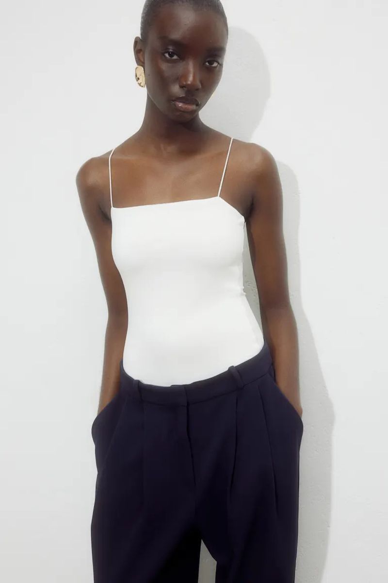 Head-and-shoulders portrait of Black female model in white spaghetti-strap top and dark trousers posing against white wall in photo studio.