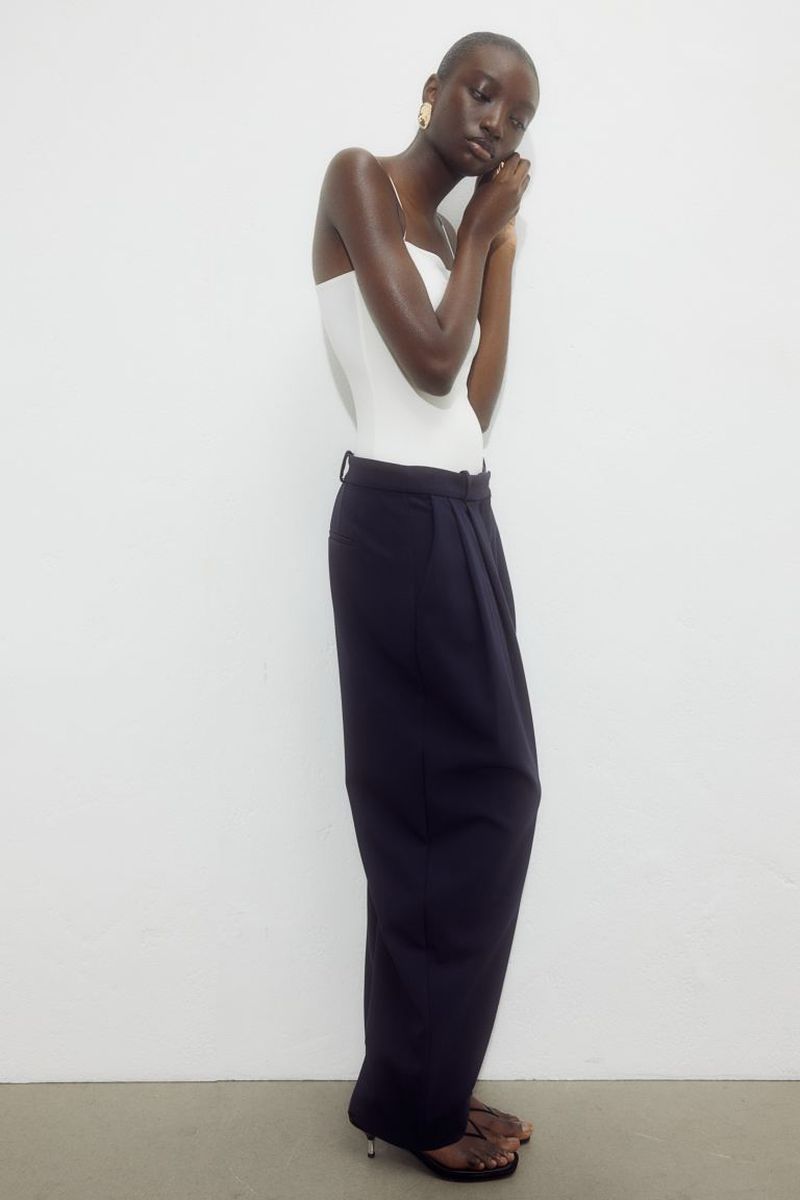 Fashion shot of a Black woman in a white top and black pants, posing in front of a white background. She is wearing gold earrings.