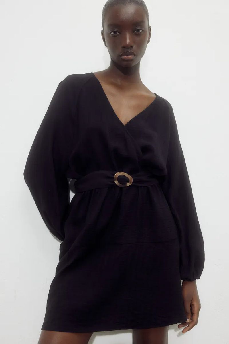 Woman in a long-sleeved black V-neck dress with a belt, posing in front of a white wall in a studio environment.