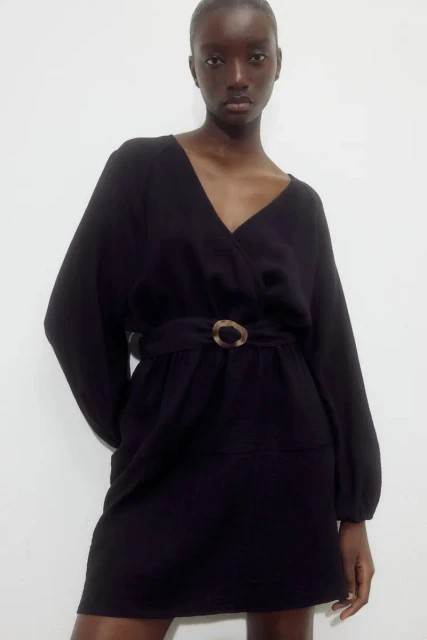 Woman in a long-sleeved black V-neck dress with a belt, posing in front of a white wall in a studio environment.