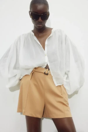 Fashion model poses in a white blouse and tan shorts with a belt. Studio shot against a white background in portrait orientation.