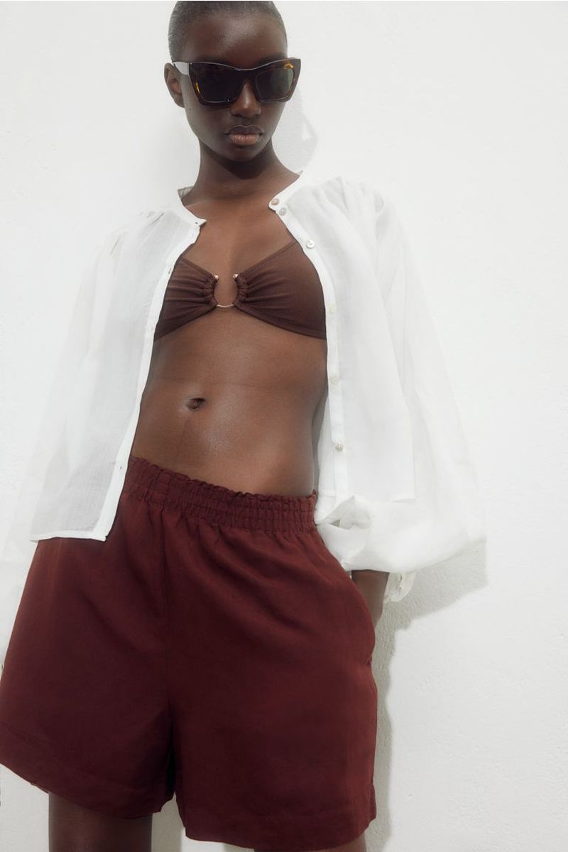 A woman wears sunglasses, a bikini top, a white button-down shirt, and dark red shorts, posing against a plain white backdrop.