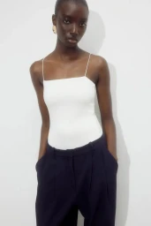 Model wearing a white spaghetti strap top and dark trousers against a white background
