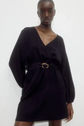 Model in a black V-neck mini dress with a round buckle belt against a plain white background for H&M