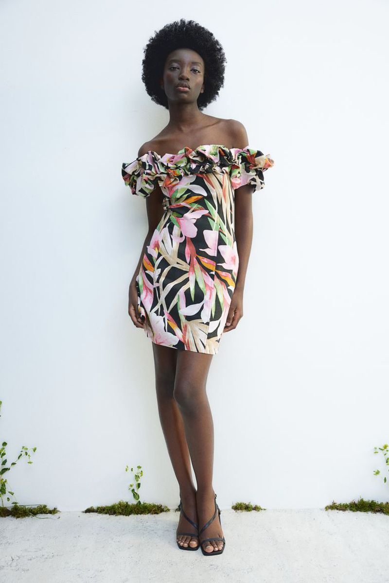 A dark-skinned woman wearing a floral print, off-the-shoulder dress and strappy heeled sandals stands against a white wall with vegetation.