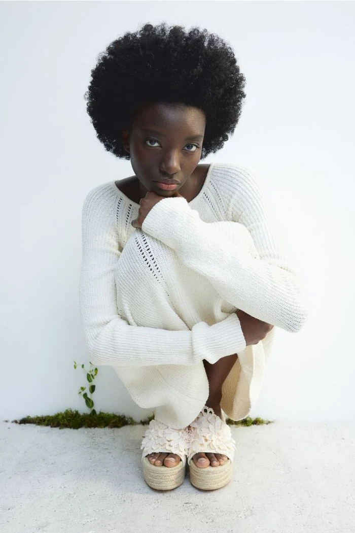 Black woman crouching against a white background wearing a white knit dress and platform sandals
