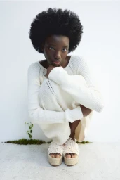 Black woman crouching against a white background wearing a white knit dress and platform sandals