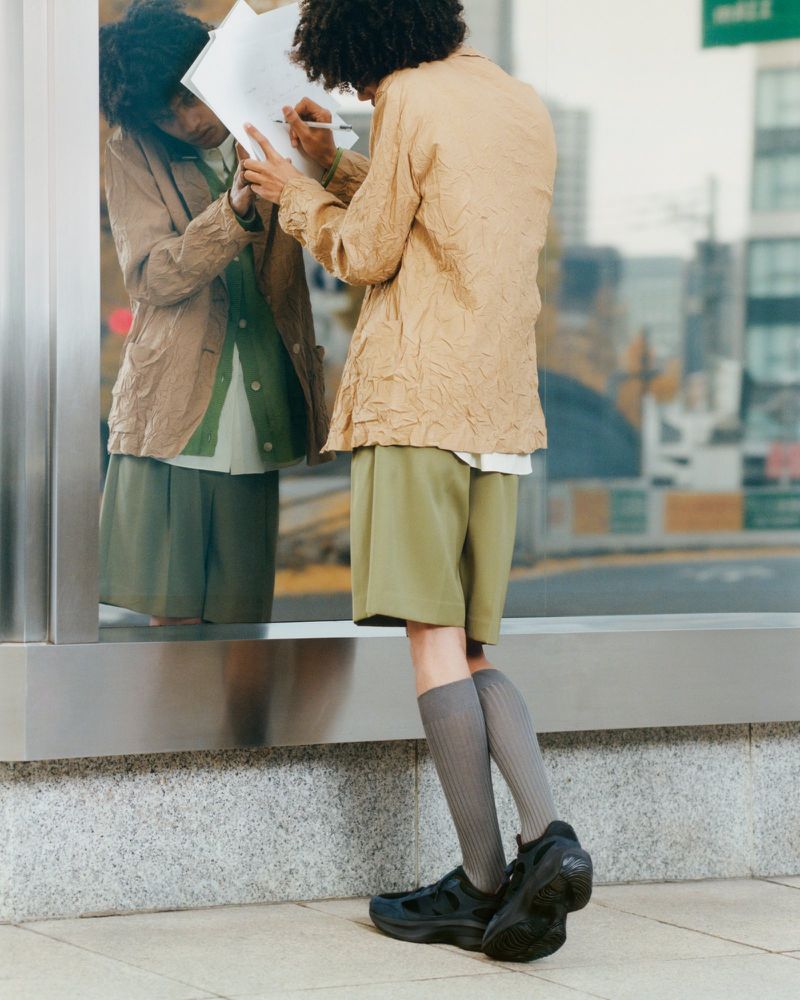 Back view of a curly-haired person in tan jacket and olive shorts standing on a city street writing on a document, reflected in glass.