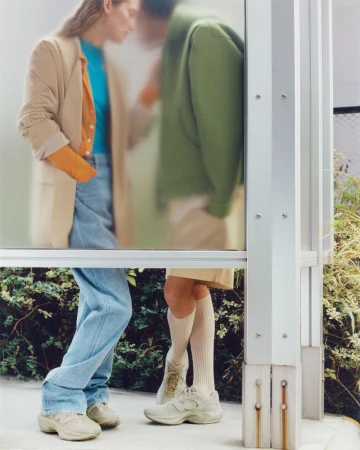 Two people stand partially visible behind a frosted glass wall, both wearing casual clothing and athletic shoes on a concrete surface.