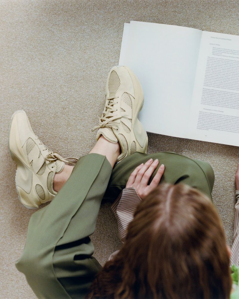 Overhead shot of a woman seated on a carpet, legs crossed, wearing sneakers and green pants, with an open book on the floor.