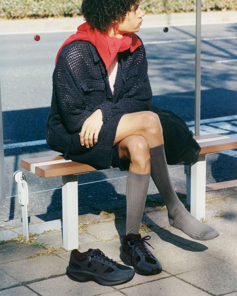 A person with curly hair wearing casual clothing sits on a bench near shoes on the ground at a bus stop in the daytime.