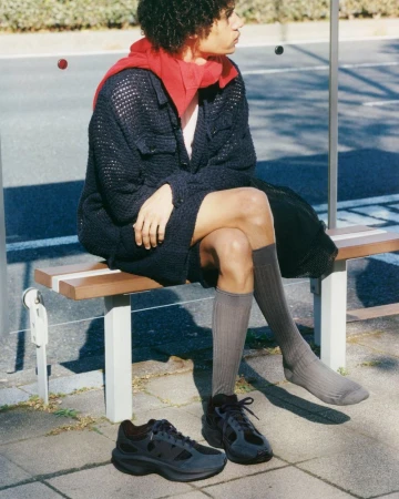A person with curly hair wearing casual clothing sits on a bench near shoes on the ground at a bus stop in the daytime.