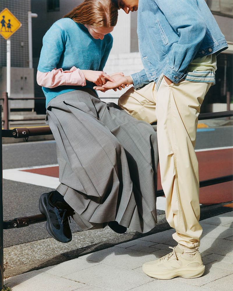 A young couple is outdoors in a city environment. The female is seated on a metal railing while the male stands facing her in an urban scene.