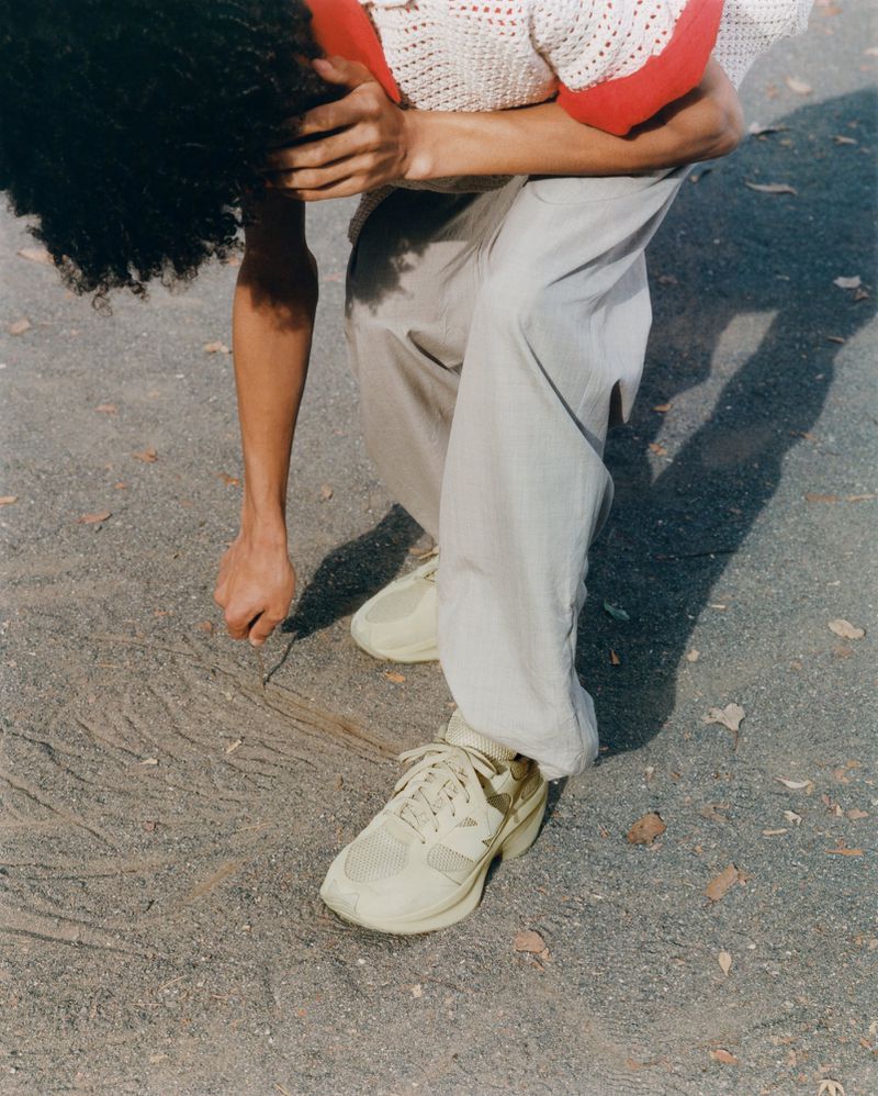 Person kneeling outdoors on gravel with running shoes and cream pants, with their head bowed and hand on the ground.