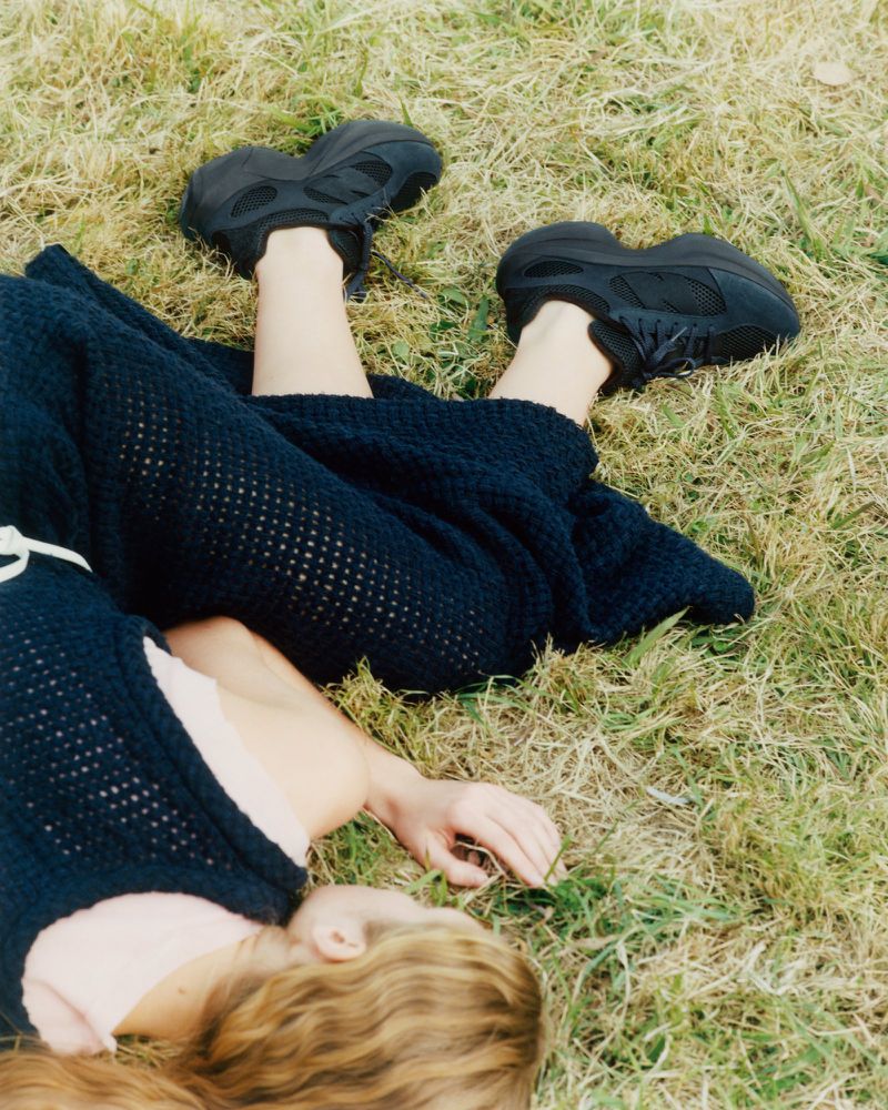 Woman lies on grass wearing a dark knit dress and black sneakers, posing with legs raised in the air in an outdoor setting.