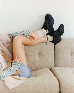 Close-up of a model wearing dark sneakers, cream socks, and striped sleeves while reclining on a textured beige sofa