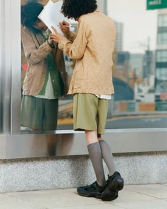 Person with curly hair wearing a tan jacket and green shorts writing on a reflective glass surface outdoors.