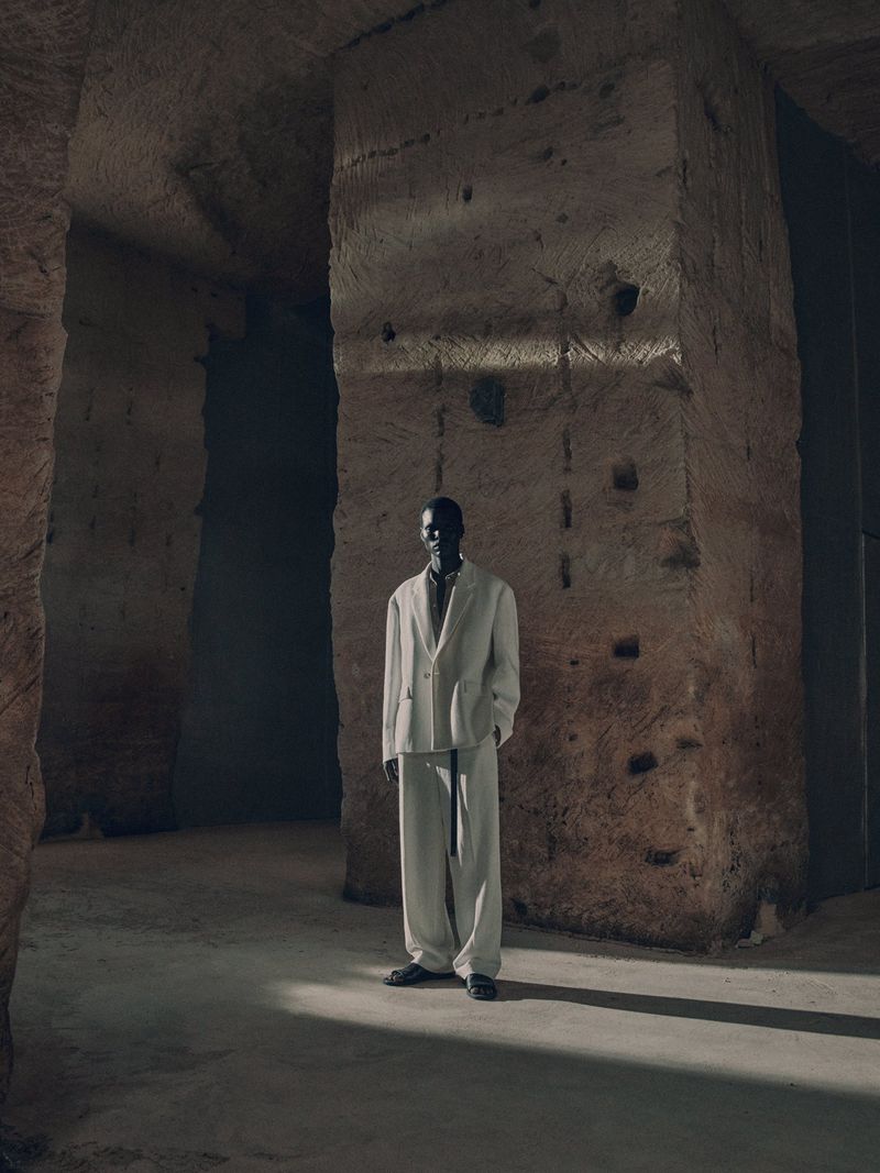 An elegant Black man in a white suit stands inside a stone tunnel, bathed in chiaroscuro lighting, in a striking fashion editorial.