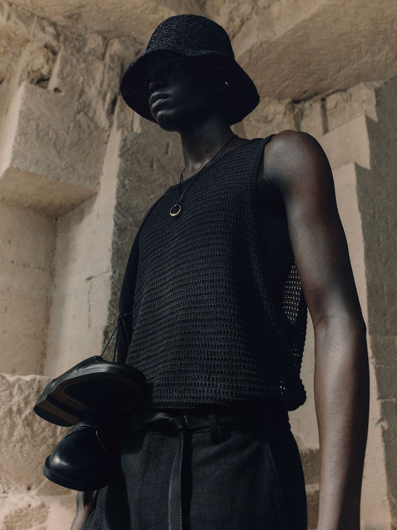 Dark-skinned male model wearing a black woven bucket hat and black shirt, holding black shoes, posing against a textured stone wall.