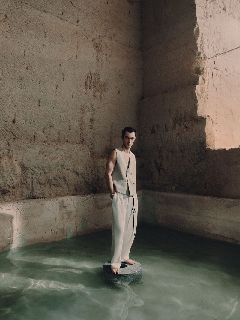 A full shot of a slender male fashion model in vest and trousers standing on a stone in water, posing in a stone room.