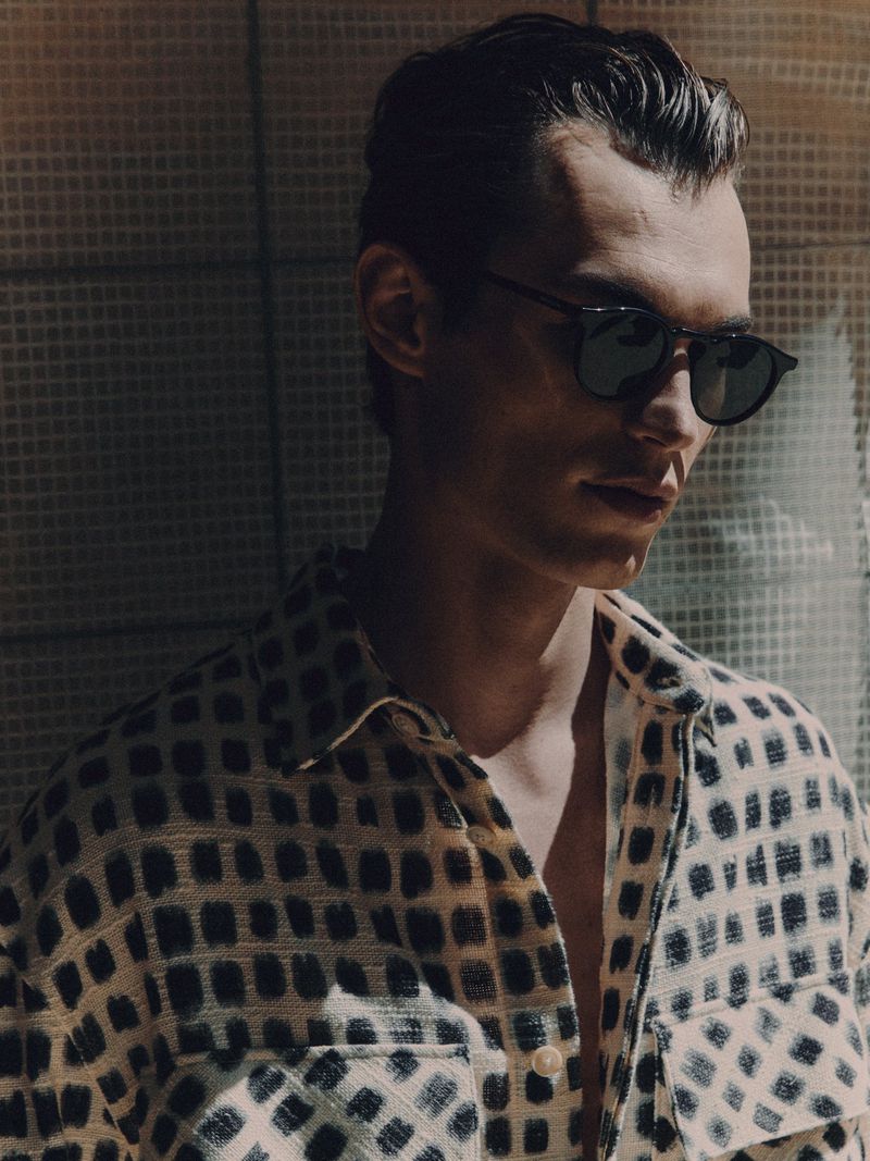 Portrait of a young adult male with sunglasses wearing an patterned shirt, posing against a textured wall in natural light.