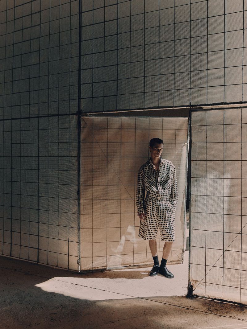 A young adult man in a patterned shirt and shorts stands in an industrial setting near a textured wall, captured in natural light.