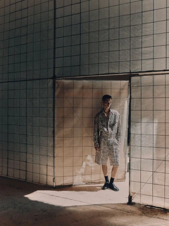 A young adult man in a patterned shirt and shorts stands in an industrial setting near a textured wall, captured in natural light.