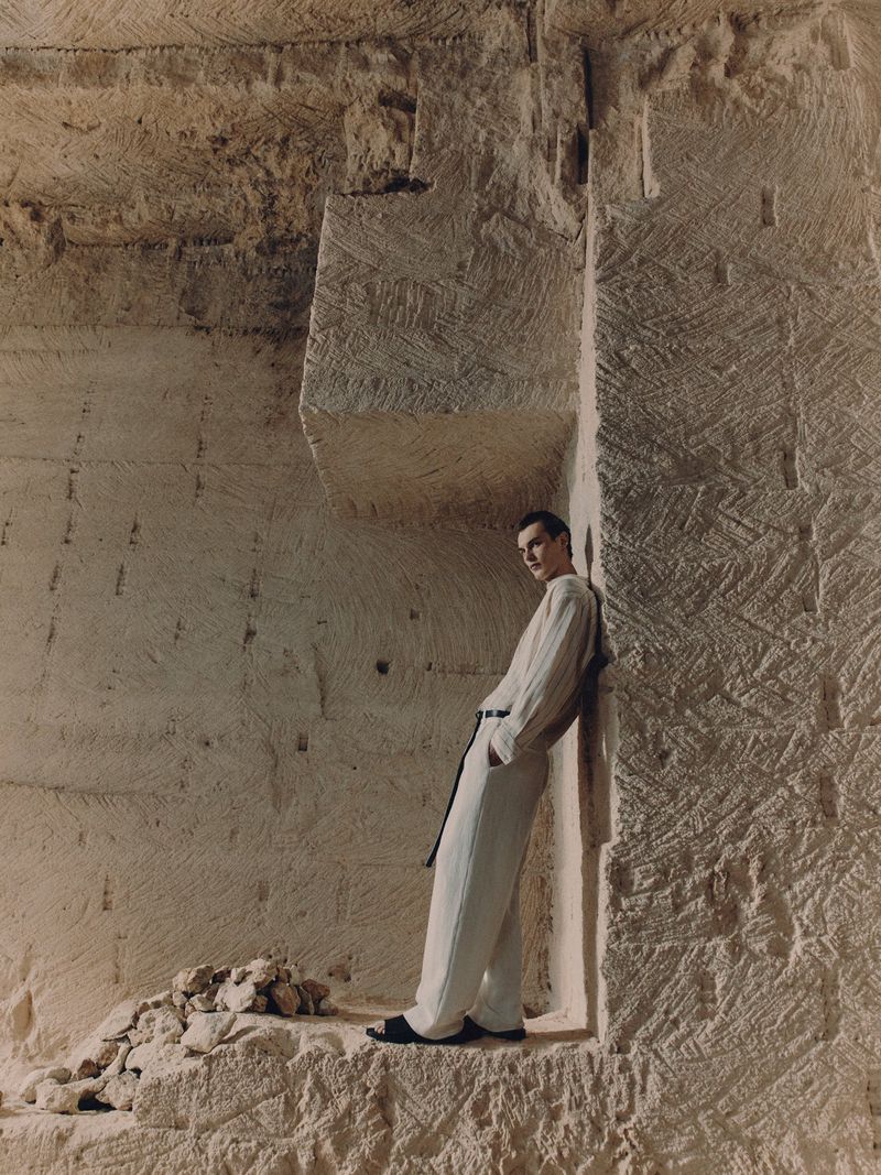 A man in a light-colored linen outfit leans against a carved stone wall in a quarry-like setting, captured in a fashion-editorial style shot.