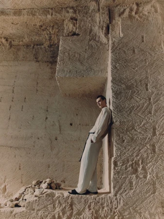 A man in a light-colored linen outfit leans against a carved stone wall in a quarry-like setting, captured in a fashion-editorial style shot.