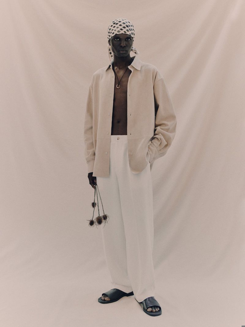 Full-length portrait of a person in a beige shirt, white pants, sandals, and headscarf, holding dried flowers against a cream backdrop.