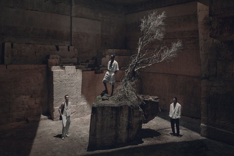 Three men pose within a stone structure with a dry tree, wearing light clothing, bathed in directed lighting with a dramatic, cavernous setting.