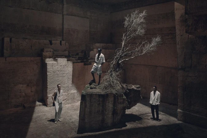 Three men pose within a stone structure with a dry tree, wearing light clothing, bathed in directed lighting with a dramatic, cavernous setting.