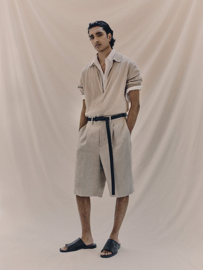 An adult male model poses in a neutral beige environment, wearing a linen outfit consisting of a short-sleeved shirt and matching shorts.