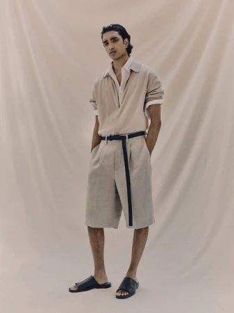 An adult male model poses in a neutral beige environment, wearing a linen outfit consisting of a short-sleeved shirt and matching shorts.