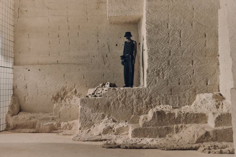 An adult stands on a stone structure against a wall in a minimalist fashion shot, conveying a sense of strength and solitude.