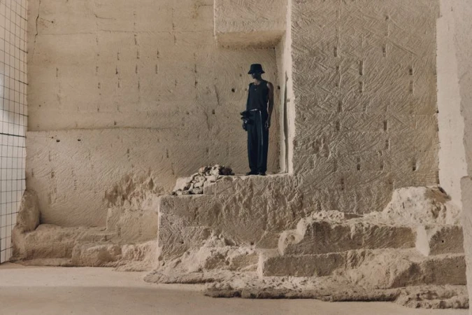 An adult stands on a stone structure against a wall in a minimalist fashion shot, conveying a sense of strength and solitude.