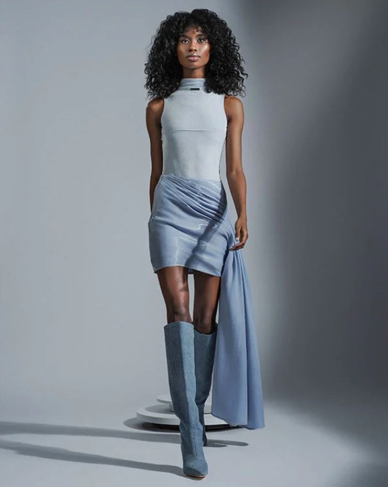 Woman in light blue sleeveless top and matching skirt with knee-high boots posing in a studio with neutral background. Fashion shot.