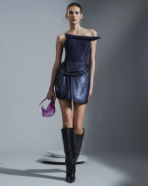 Model Elodie Safeno wearing a structured dark blue dress and black boots, holding a purple handbag against a grey backdrop.