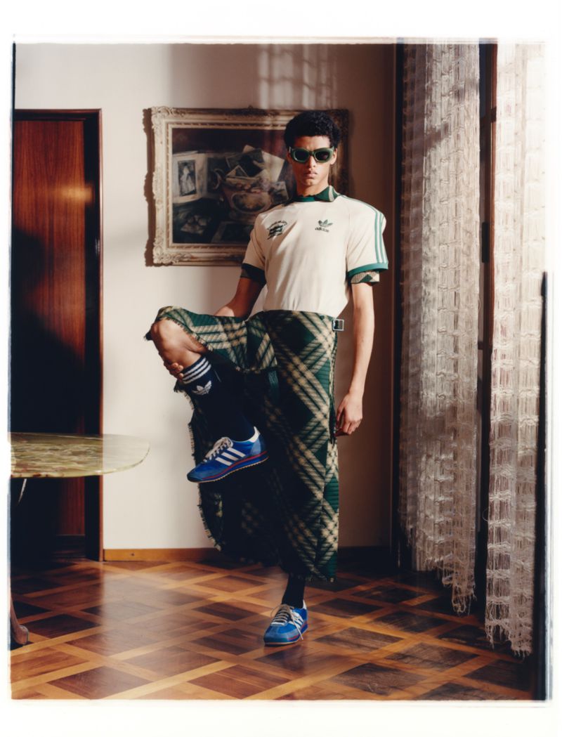 A young adult man strikes a pose indoors, wearing a cream polo shirt with green accents, green plaid pants, and blue Adidas sneakers.
