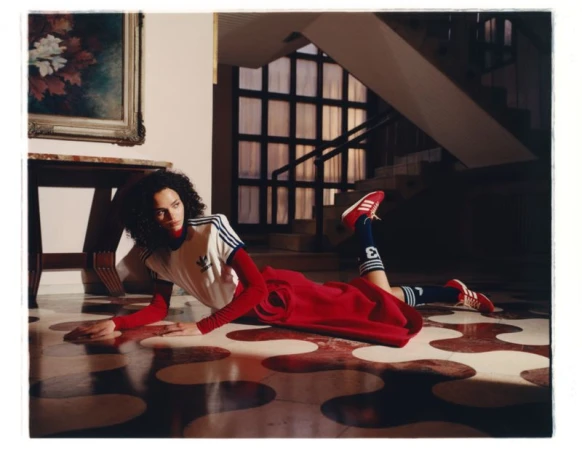 Fashion shot of a young adult woman in sportswear lying on a patterned floor with a staircase and framed painting in the background.